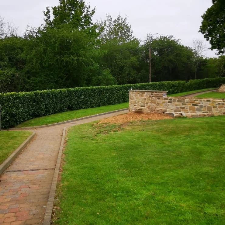 Brick walkway curves through a green lawn past a stone wall and a tall manicured hedge.